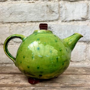 May include: A round, lime green ceramic teapot with a glossy finish and a subtle crackle texture. The teapot features small, raised green dots across its surface, a curved handle, a pointed spout, and a dark brown lid knob and base feet.