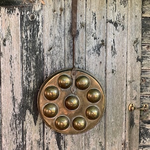 Peut inclure: Une poêle à escargots en laiton avec neuf cavités rondes, suspendue à une porte en bois vieilli. La poêle, avec une longue poignée, présente une patine ancienne, témoignant de son utilisation et de son âge.