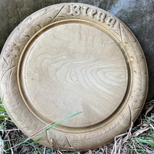 May include: A round, light-colored wooden serving tray with carved floral and leaf designs around the rim. The word "Bread" is carved into the top edge. The tray is sitting on grass.