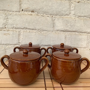 May include: Four brown ceramic soup bowls with lids and handles. The bowls are glossy and have a rounded shape. The lids have a small knob on top. The bowls are arranged on a wooden surface against a white brick wall.