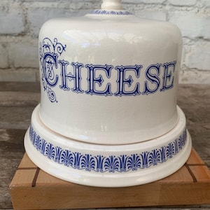 Könnte beinhalten: Weißer Keramik-Käseglocke mit blauem "CHEESE"-Schriftzug und dekorativem blauen Rand. Die Haube hat einen Griff und steht auf einem passenden Sockel. Die Käseglocke steht auf einem Holzbrett.