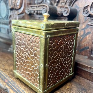 May include: A square box with a lid featuring a brass frame and a small brass knob handle. The four sides of the box are made of textured copper panels with a pebble-like pattern. The brass frame is secured with small rivets at each corner.