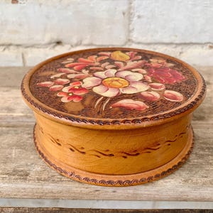 以下が含まれることがあります： 蓋に花の模様が施された丸い木製の箱。蓋には、赤、ピンク、黄色の花が詳細に彫刻されています。箱は明るい茶色の仕上げで、木製の表面に置かれています。