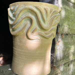 May include: A handcrafted, beige ceramic vase with a textured, wavy design around the rim. The vase has a cylindrical shape and sits on a wooden surface. The upper portion features a light green and beige swirl pattern.