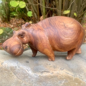 Könnte beinhalten: Eine Holzskulptur eines Nilpferds. Das Nilpferd ist aus hellbraunem Holz mit dunkleren Maserungsmustern geschnitzt. Die Skulptur hat einen abgerundeten Körper, kurze Beine und eine große Schnauze. Das Nilpferd ist in einer stehenden Position dargestellt.