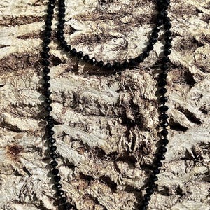 Puede incluir: Un collar largo de cuentas negras descansa sobre un fondo marr&oacute;n texturizado. El collar est&aacute; compuesto por peque&ntilde;as cuentas redondas, creando un dise&ntilde;o simple pero elegante. Las cuentas capturan la luz, a&ntilde;adiendo un brillo sutil.