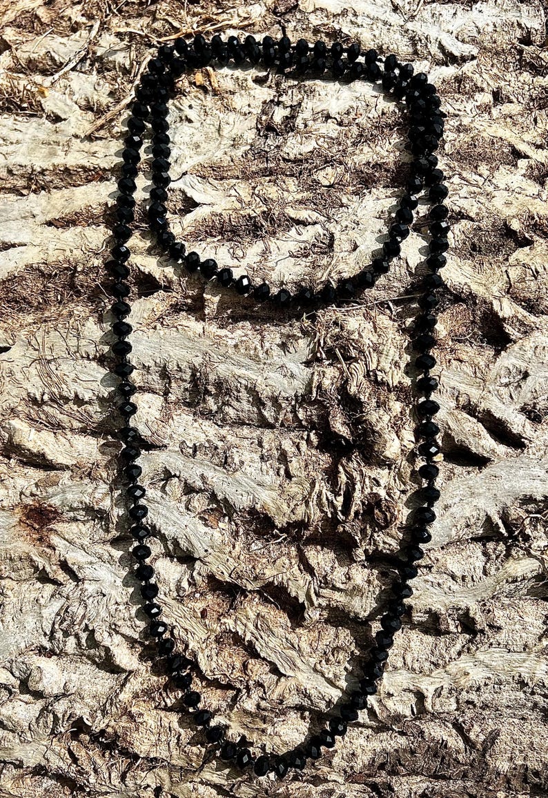 Puede incluir: Un collar largo de cuentas negras descansa sobre un fondo marr&oacute;n texturizado. El collar est&aacute; compuesto por peque&ntilde;as cuentas redondas, creando un dise&ntilde;o simple pero elegante. Las cuentas capturan la luz, a&ntilde;adiendo un brillo sutil.