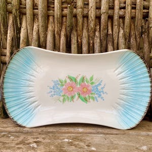 Könnte beinhalten: Eine weiße Keramik-Servierplatte mit einem blauen und weißen gewellten Rand. Die Platte zeigt ein Blumenmuster mit rosa Blüten und grünen Blättern.
