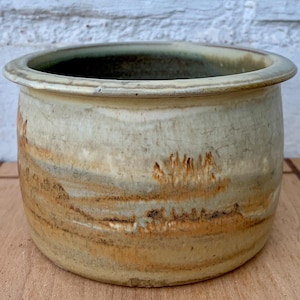 May include: A handcrafted ceramic pot with a wide, rounded shape. The exterior features a blend of earthy tones, including beige, brown, and hints of green. The rim is slightly flared, and the pot has a rustic, textured appearance, suggesting a natural, organic aesthetic.