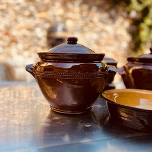 May include: A dark brown glazed ceramic pot with a matching lid and two side handles. The pot features a decorative embossed band of small circular patterns around the upper body. The surface is smooth and reflective with a deep, glossy finish.
