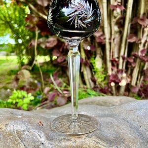 Könnte beinhalten: Ein Kristall-Weinglas mit einer tief burgunderfarbenen Schale und einem klaren, facettierten Stiel. Die Schale hat ein Sternmuster. Das Glas steht auf einer grauen Steinoberfläche, mit einem verschwommenen grünen und violetten Hintergrund.