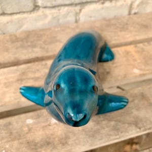 May include: A teal ceramic seal figurine with a glossy finish. The seal is lying down with its flippers extended. The figurine has dark brown eyes and is placed on a wooden surface.