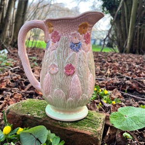 Puede incluir: Jarra de cerámica con un esmalte rosa claro, adornada con diseños florales en relieve en tonos rosa, amarillo y azul. La jarra tiene un asa curva y un pico ancho. La base es de color verde claro.