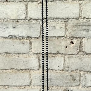 Puede incluir: Un collar largo de cuentas negras cuelga verticalmente contra una pared de ladrillos blancos texturizados. Las cuentas son peque&ntilde;as y est&aacute;n espaciadas uniformemente, creando un dise&ntilde;o simple y elegante. El collar es un accesorio vers&aacute;til.