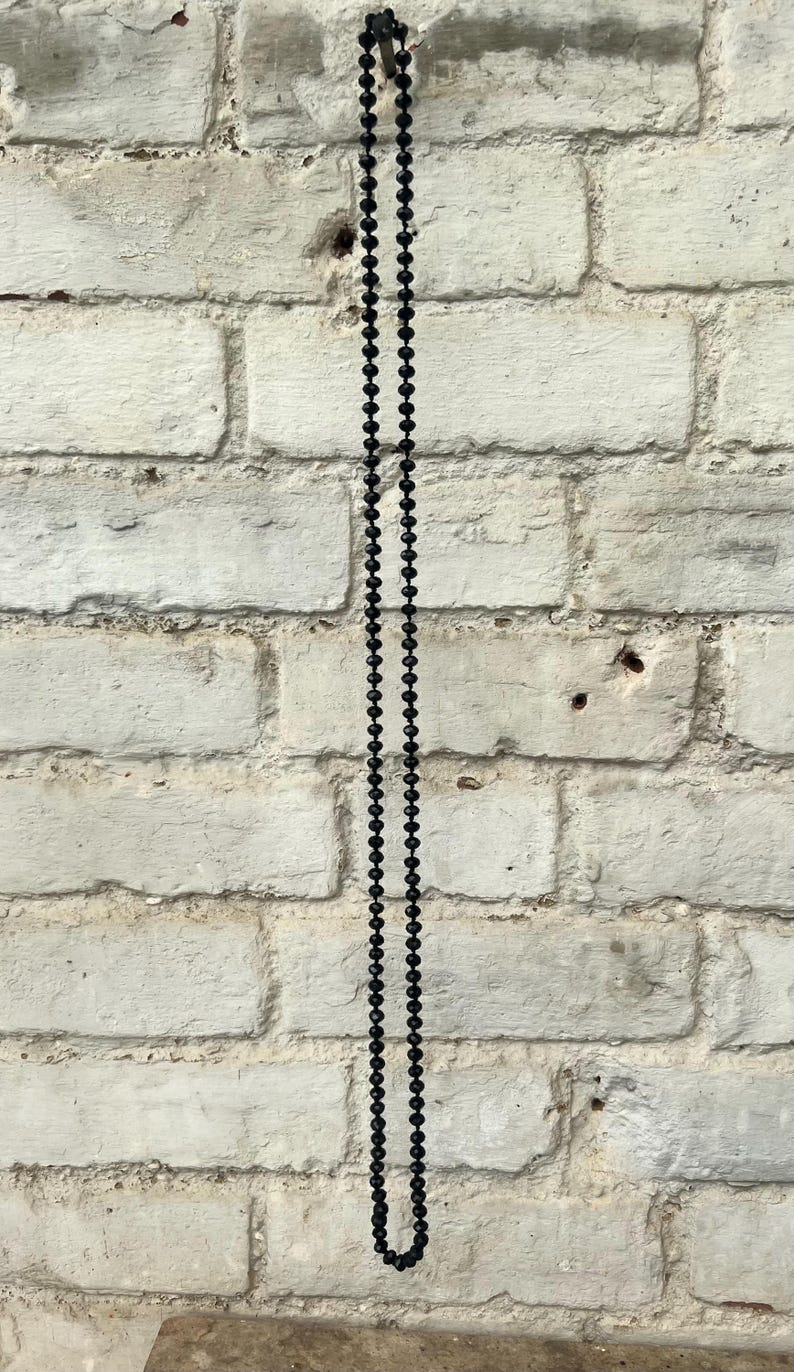 Puede incluir: Un collar largo de cuentas negras cuelga verticalmente contra una pared de ladrillos blancos texturizados. Las cuentas son peque&ntilde;as y est&aacute;n espaciadas uniformemente, creando un dise&ntilde;o simple y elegante. El collar es un accesorio vers&aacute;til.