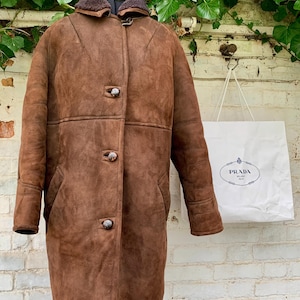 Puede incluir: Un abrigo de piel de oveja marrón con cuello de piel y tres botones. El abrigo está en un maniquí y hay una bolsa de compras blanca con el logotipo de Prada en el fondo.