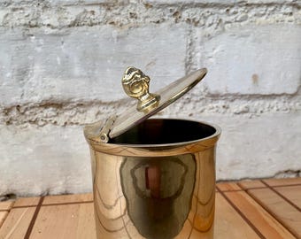 Peut inclure: Un récipient cylindrique en laiton poli avec un couvercle à charnière. Le couvercle est doté d'une petite poignée en forme de bouton sculpté en laiton. Le récipient présente une surface métallique lisse et réfléchissante avec une base circulaire.