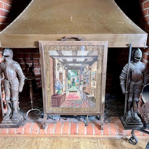 May include: A rectangular wooden fireplace screen featuring a detailed needlepoint tapestry of an indoor hallway leading to a garden view. The scene includes a floral arrangement, a shelf with blue plates, and a red rug on a tiled floor.