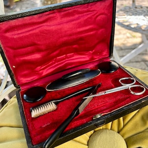 May include: A black rectangular case with a vibrant red satin interior lining containing a set of grooming tools. The set includes a small black brush with light bristles, a pair of silver metal scissors, and several black handled implements.