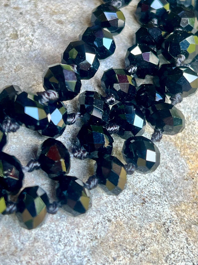 Puede incluir: Primer plano de un collar de cuentas negras. Las cuentas est&aacute;n facetadas, reflejando la luz, y ensartadas con hilo oscuro. El collar descansa sobre una superficie texturizada de color claro.