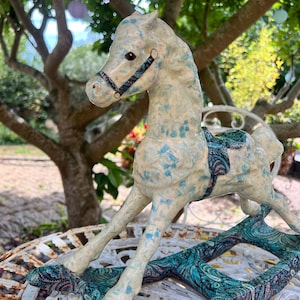 May include: A ceramic rocking horse figurine featuring a cream-colored body with a speckled blue and pink pattern. The horse has a dark bridle and a saddle, and it is mounted on curved rockers decorated with a detailed teal and purple paisley print.