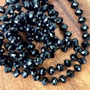 Puede incluir: Un collar largo de cuentas negras con cuentas facetadas. Las cuentas est&aacute;n ensartadas en un cord&oacute;n negro y est&aacute;n dispuestas en un patr&oacute;n en capas. El collar se exhibe sobre una superficie de madera.