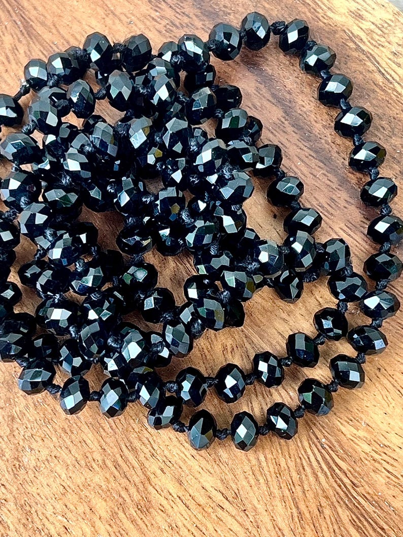 Puede incluir: Un collar largo de cuentas negras con cuentas facetadas. Las cuentas est&aacute;n ensartadas en un cord&oacute;n negro y est&aacute;n dispuestas en un patr&oacute;n en capas. El collar se exhibe sobre una superficie de madera.