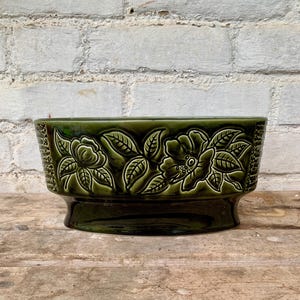 May include: A dark green ceramic planter with a raised floral design. The rectangular planter has a glossy finish and a pedestal base. The embossed flowers and leaves add a decorative touch to this vintage-style container.