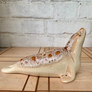 May include: A ceramic seal ornament in a resting pose. The seal is mainly beige with brown spotted detailing on its back. The figurine has a smooth, glossy finish and is set against a light-coloured background.