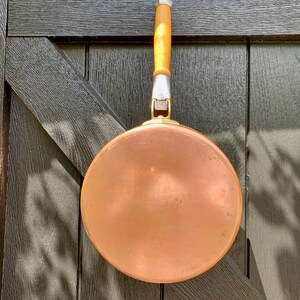 May include: A copper frying pan with a wooden handle and metal detailing. The pan is round and hangs against a dark wooden background. The handle is light brown and silver-toned.