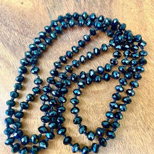 Puede incluir: Un collar largo de cuentas facetadas de color azul oscuro. Las cuentas est&aacute;n ensartadas, creando un efecto de capas. El collar se muestra sobre una superficie de madera, mostrando su color y dise&ntilde;o.