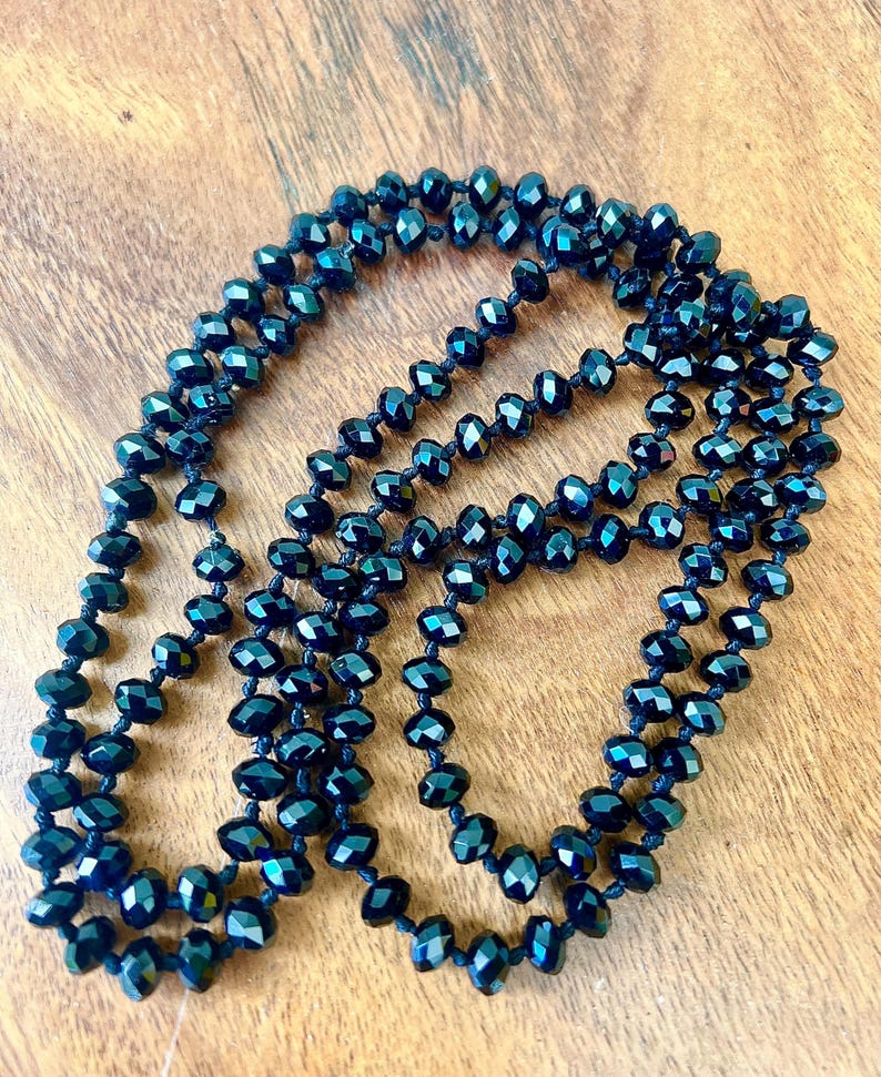 Puede incluir: Un collar largo de cuentas facetadas de color azul oscuro. Las cuentas est&aacute;n ensartadas, creando un efecto de capas. El collar se muestra sobre una superficie de madera, mostrando su color y dise&ntilde;o.