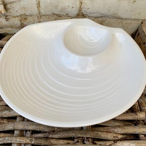 Puede incluir: Plato de servir de cerámica blanca con un bol central para salsas. El plato tiene un borde festoneado y una superficie texturizada y ondulada.