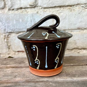 May include: A dark brown ceramic sugar bowl with a matching lid featuring a curved handle. The exterior is decorated with vertical light blue and cream-colored abstract patterns. The base of the bowl is unglazed, revealing a natural terracotta color.