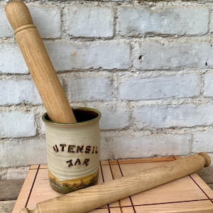 Könnte beinhalten: Ein Keramik-Utensilienbehälter mit den Worten "UTENSIL JAR" in geprägten Buchstaben. Ein Nudelholz aus Holz befindet sich im Inneren des Behälters, und ein weiteres Nudelholz liegt auf einem Holzschneidebrett. Der Behälter hat eine beige Basis mit einem grünen und gelben Landschaftsdesign.