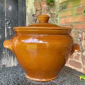 May include: A brown ceramic crock with a lid. The crock has two handles on the sides and a rounded shape.