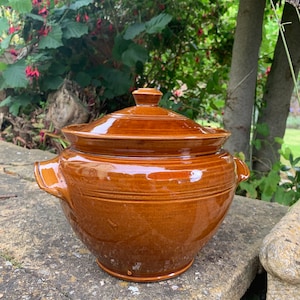 May include: A brown ceramic pot with a lid and two handles. The pot has a glossy finish and is sitting on a stone surface.