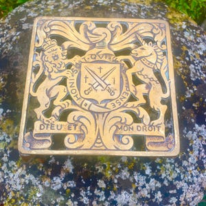Puede incluir: Un cuadrado de metal dorado con un escudo de armas. El escudo de armas presenta un león y un unicornio, un escudo con espadas cruzadas y el texto "Dieu et Mon Droit".