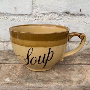 Puede incluir: Una taza de sopa de cerámica marrón y beige con asa. La taza tiene la palabra "Soup" escrita en cursiva negra en la parte delantera.