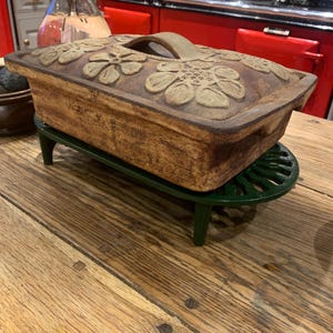 May include: A brown ceramic baking dish with a floral design and a handle. The dish is sitting on a green metal stand.