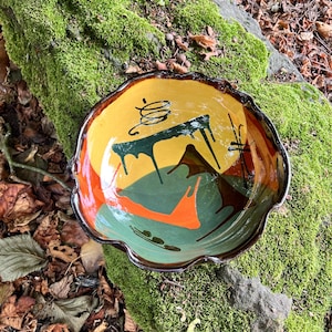 Pode incluir: Uma tigela de cerâmica com borda recortada e acabamento brilhante. O interior apresenta um design abstrato pintado à mão com seções ousadas em amarelo mostarda, verde sálvia e laranja brilhante, acentuado por padrões de gotas castanhas.