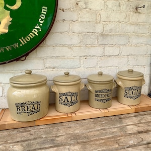 Può includere: Quattro barattoli da cucina in ceramica beige con coperchi, etichettati con "BREAD", "SALT", "DRIED FRUIT" e "SUGAR" in testo nero. I barattoli sono disposti su un vassoio di legno contro un muro di mattoni bianchi. I barattoli hanno un'estetica vintage.