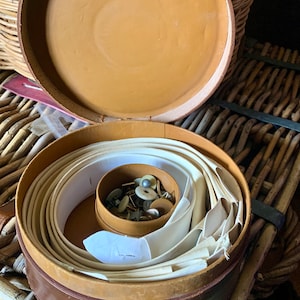 Puede incluir: Una caja de cuero marrón con la tapa abierta, que revela una cinta de tela blanca enrollada alrededor de una caja marrón más pequeña. La caja más pequeña contiene una variedad de pequeños botones de metal y plástico.