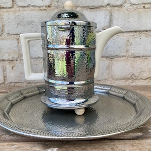Könnte beinhalten: Eine Vintage-Kaffeekanne aus Chrom mit gehämmertem Muster, mit weißem Griff, Ausguss und Knauf. Die Kanne steht auf einem passenden Tablett aus gehämmertem Metall. Das Design erinnert an Küchenutensilien der Moderne aus der Mitte des Jahrhunderts.