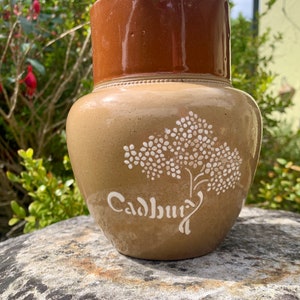 以下が含まれることがあります： 白い花柄と白い文字で「Cadbury」と書かれた茶色の陶器のピッチャー