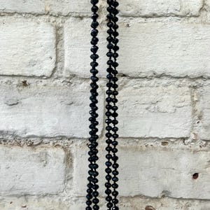 Puede incluir: Un collar largo de cuentas negras cuelga contra una pared de ladrillos blancos. El collar est&aacute; compuesto por muchas cuentas peque&ntilde;as y facetadas, creando un accesorio oscuro y elegante. Las cuentas reflejan la luz.