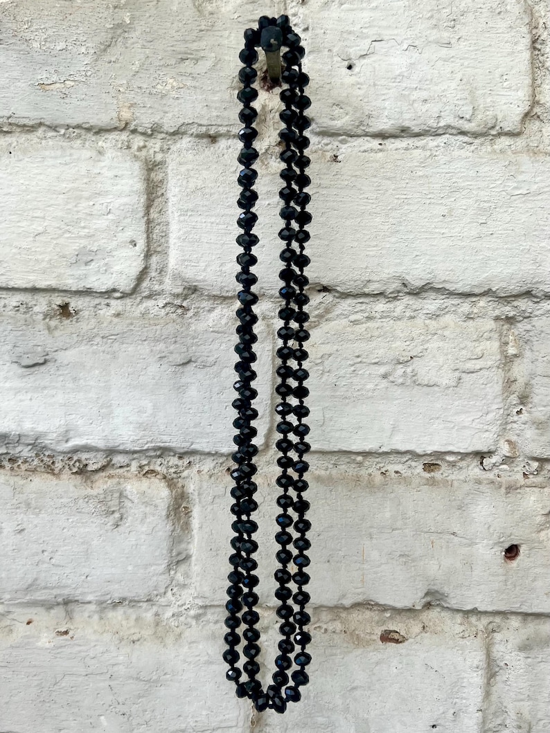 Puede incluir: Un collar largo de cuentas negras cuelga contra una pared de ladrillos blancos. El collar est&aacute; compuesto por muchas cuentas peque&ntilde;as y facetadas, creando un accesorio oscuro y elegante. Las cuentas reflejan la luz.