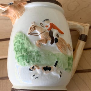 Puede incluir: Jarra de cerámica color crema con un pico en forma de cabeza de zorro y un asa marrón. La jarra presenta una escena en relieve de un jinete a caballo saltando una valla, con un perro corriendo debajo. La base y el borde son marrones.
