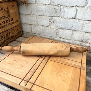 Può includere: Un mattarello in legno color marrone chiaro con un corpo cilindrico e manici affusolati su ciascuna estremità. Il legno presenta una venatura naturale con segni visibili di usura e consistenza su tutta la superficie di questo utensile da cucina.