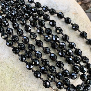 Puede incluir: Collar de varias hebras con cuentas negras facetadas ensartadas en un cord&oacute;n negro. Las cuentas tienen un acabado brillante y est&aacute;n conectadas con un cierre plateado. El collar se exhibe sobre una superficie de piedra clara.